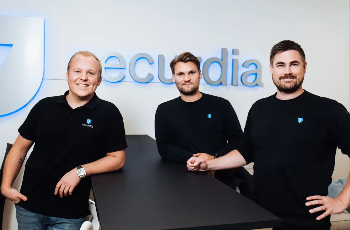 Tre män poserar framför en vägg med texten "securdia", alla bär svarta tröjor med företagets logotyp.