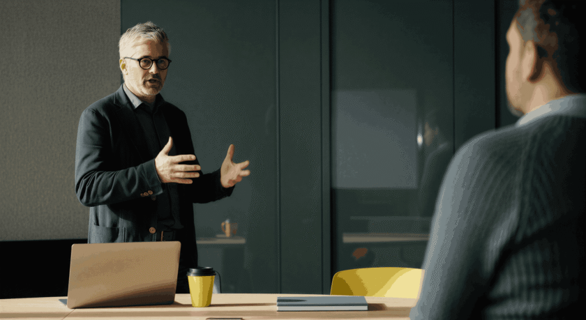 Äldre man i kostym gestikulerar vid ett skrivbord med laptop och kaffemugg, samtalar med en annan person.