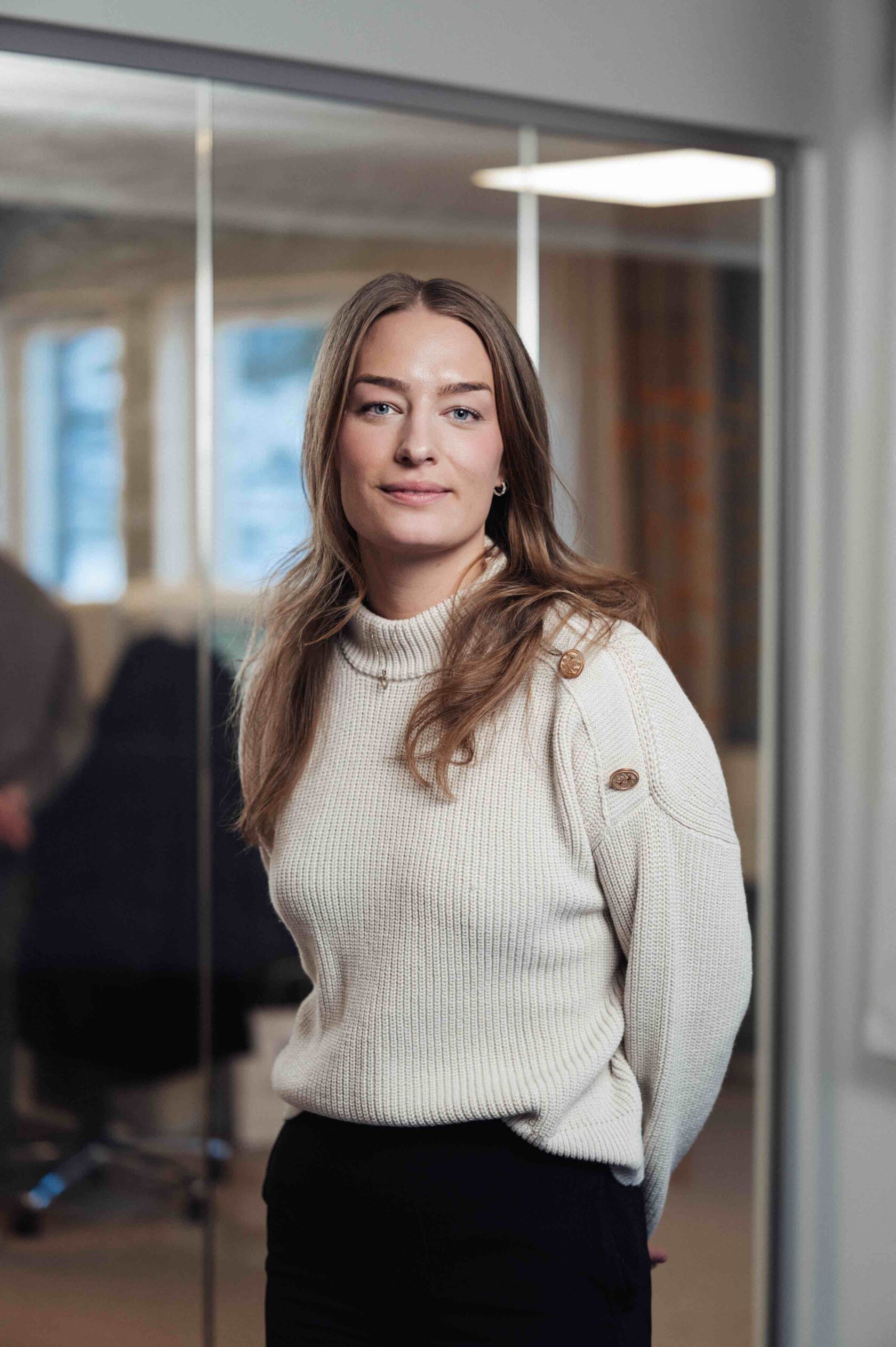 Kvinna med långt brunt hår i vit stickad tröja står i ett kontorsrum med glasväggar i bakgrunden.