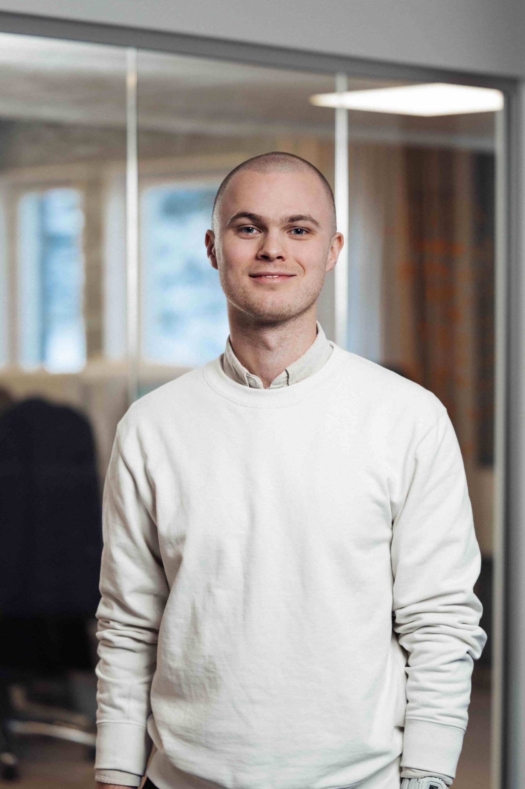Ung man med rakat huvud och vit tröja står leende inomhus framför glasvägg på kontor.