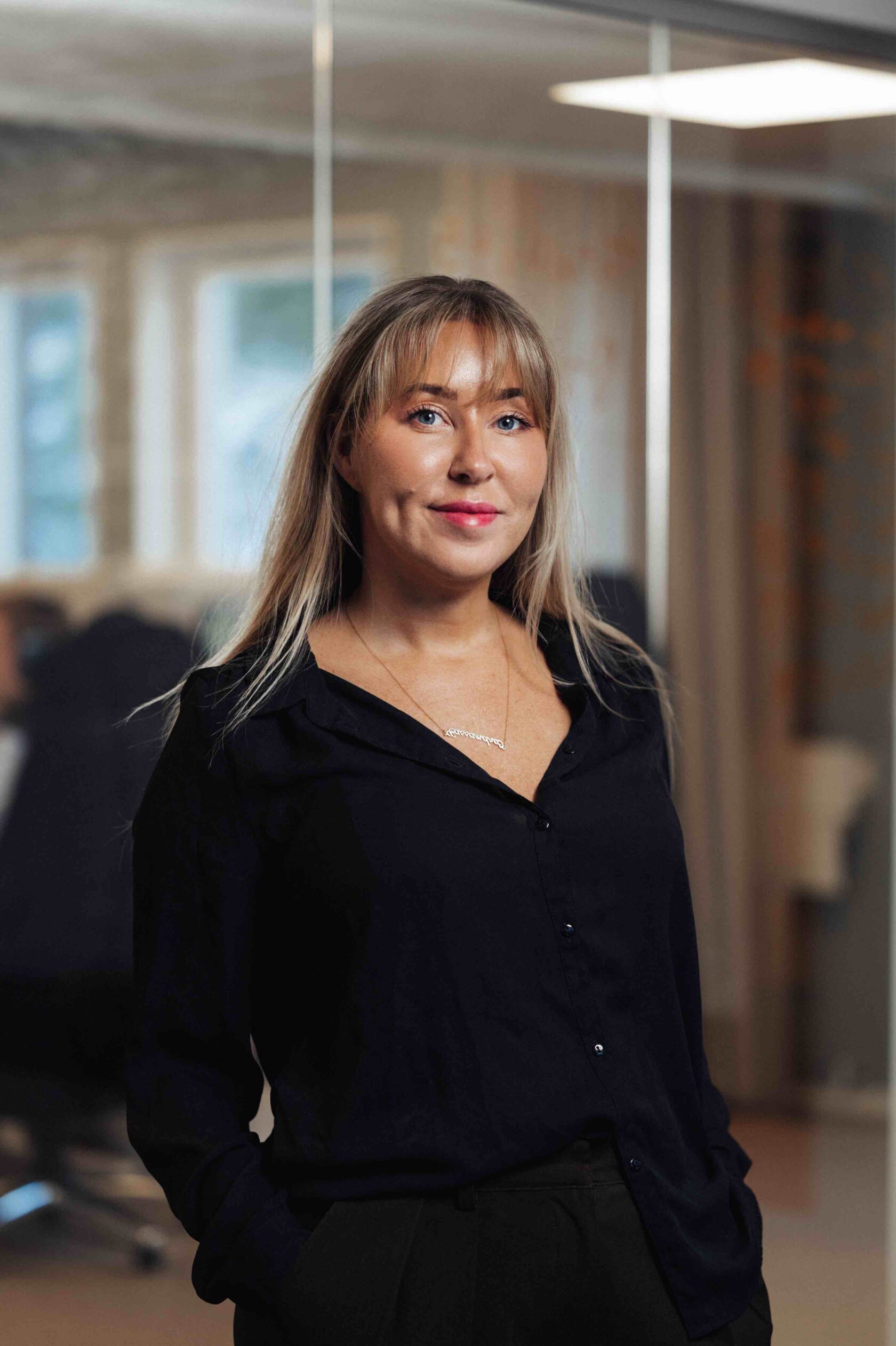Kvinna med långt blont hår och svart skjorta står i ett modernt kontorsrum och ler mot kameran.