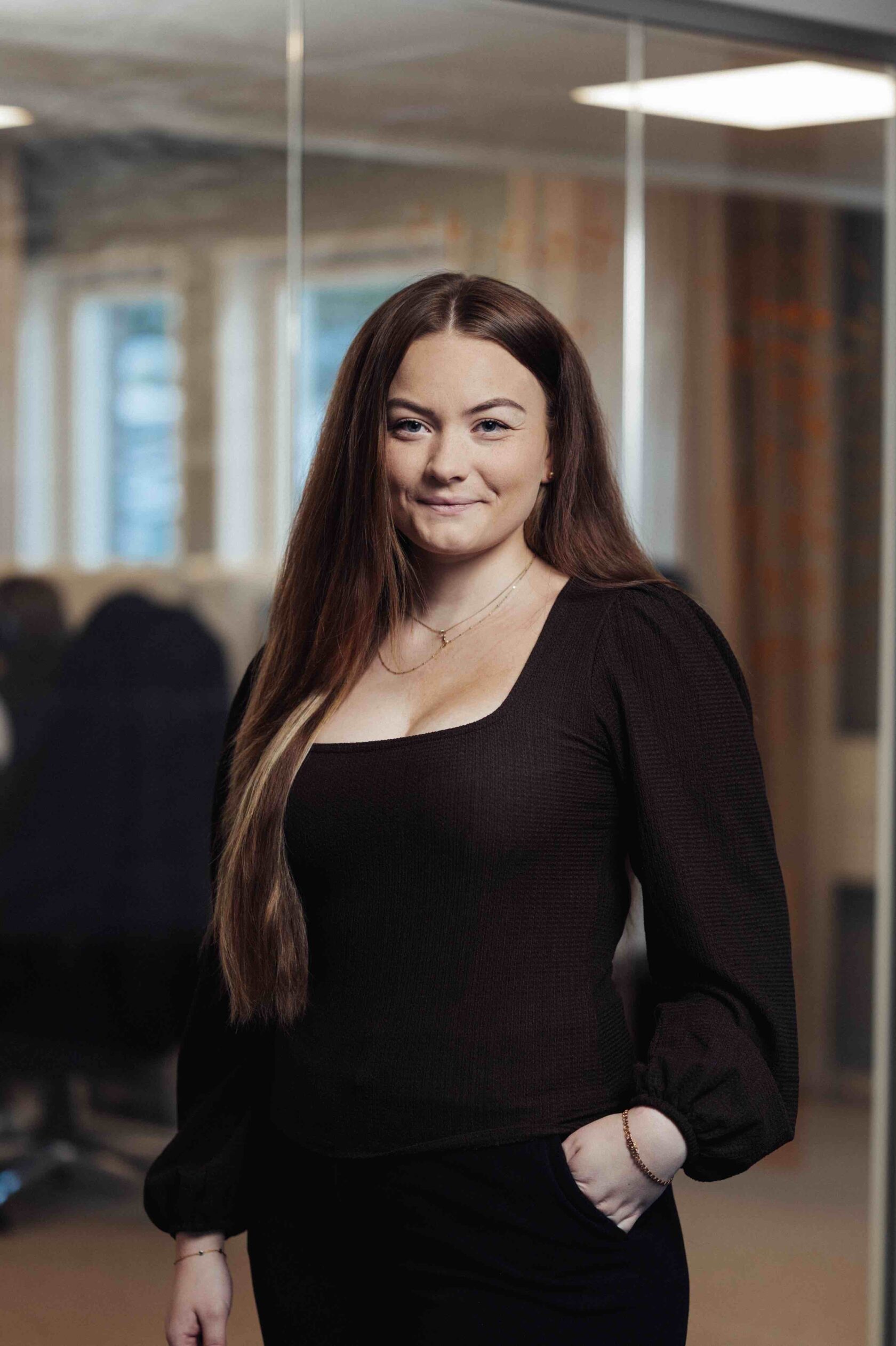 Kvinna med långt brunt hår i svart tröja står i ett kontor och ler mot kameran.